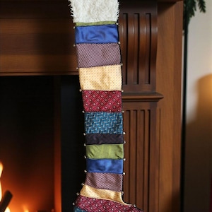 May include: A handmade Christmas stocking made from repurposed neckties. The stocking features a white fluffy cuff and is constructed from various patterned fabrics in shades of blue, red, yellow, and green. Silver studs line the sides.