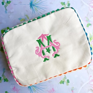 Puede incluir: Bolsa de cosméticos rectangular color crema con un monograma bordado en rosa y verde. La bolsa tiene un borde festoneado en verde, azul, naranja y rosa. El fondo es un patrón colorido.