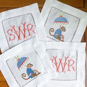 May include: Four white linen cocktail napkins. Two napkins feature coral-colored monogrammed initials. The other two napkins have an embroidered monkey wearing a blue coat and holding a blue umbrella with red and green trim.