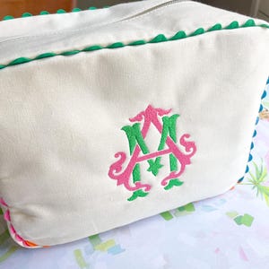 Puede incluir: Bolsa de cosm&eacute;ticos rectangular de color crema con cierre de cremallera y ribete festoneado verde. La bolsa presenta un monograma bordado en rosa y verde. La bolsa est&aacute; hecha de un material similar a la lona.