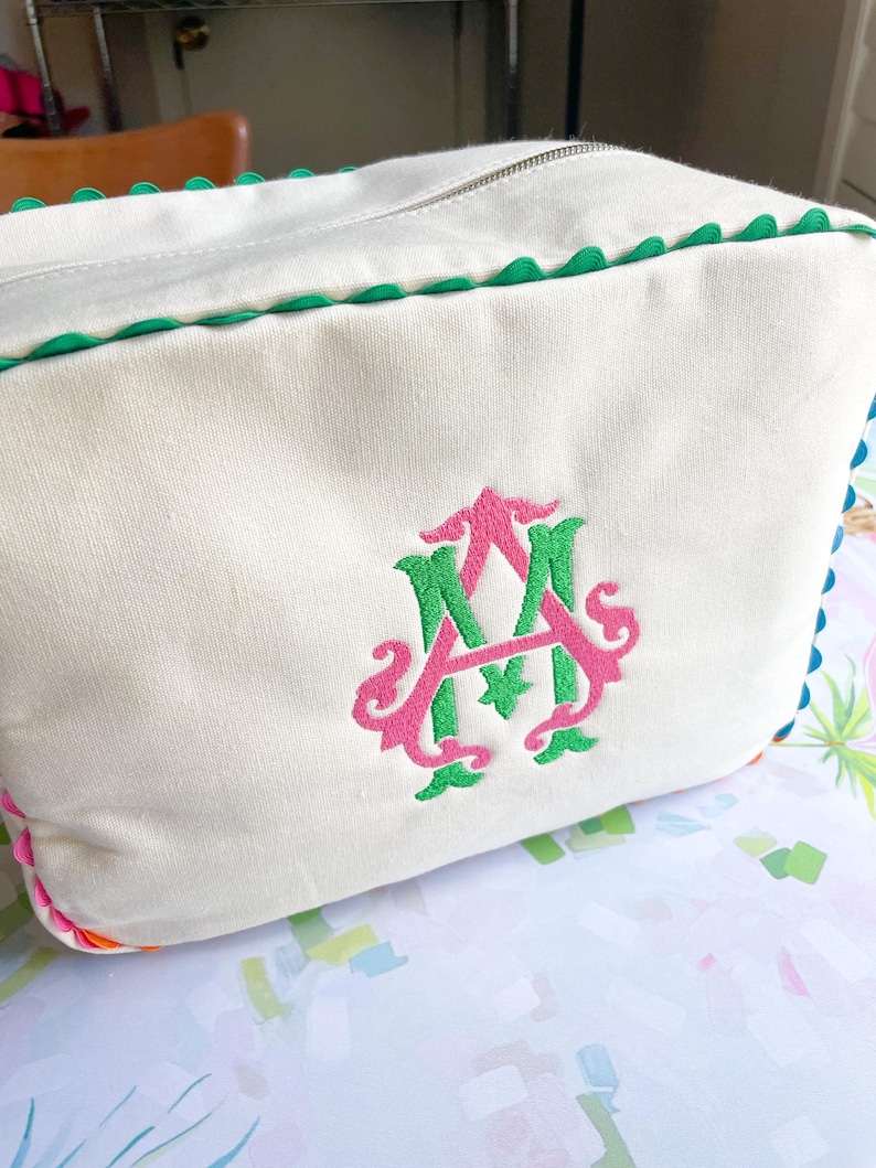 Puede incluir: Bolsa de cosm&eacute;ticos rectangular de color crema con cierre de cremallera y ribete festoneado verde. La bolsa presenta un monograma bordado en rosa y verde. La bolsa est&aacute; hecha de un material similar a la lona.