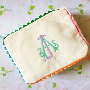 Puede incluir: Bolsa rectangular color crema con un borde festoneado colorido. Los bordes est&aacute;n adornados con verde, rosa, naranja y azul. Un monograma bordado en tonos morados y verdes est&aacute; centrado en la parte delantera.