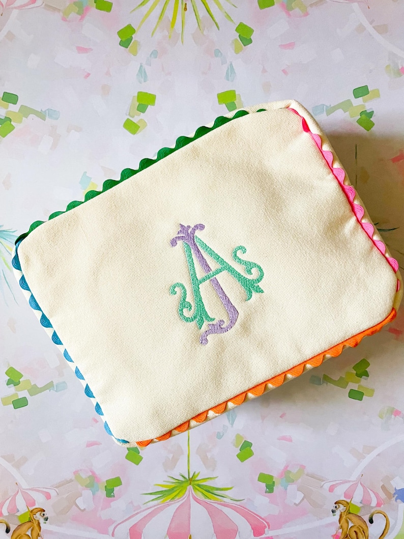 Puede incluir: Bolsa rectangular color crema con un borde festoneado colorido. Los bordes est&aacute;n adornados con verde, rosa, naranja y azul. Un monograma bordado en tonos morados y verdes est&aacute; centrado en la parte delantera.