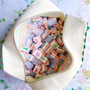 Puede incluir: Una bolsa de lona blanca, abierta y llena de coloridas fichas de Mahjong. Las fichas tienen una mezcla de colores, incluyendo naranja, lavanda y blanco. La bolsa tiene un ribete verde y una cremallera dorada. El fondo tiene un patr&oacute;n caprichoso.