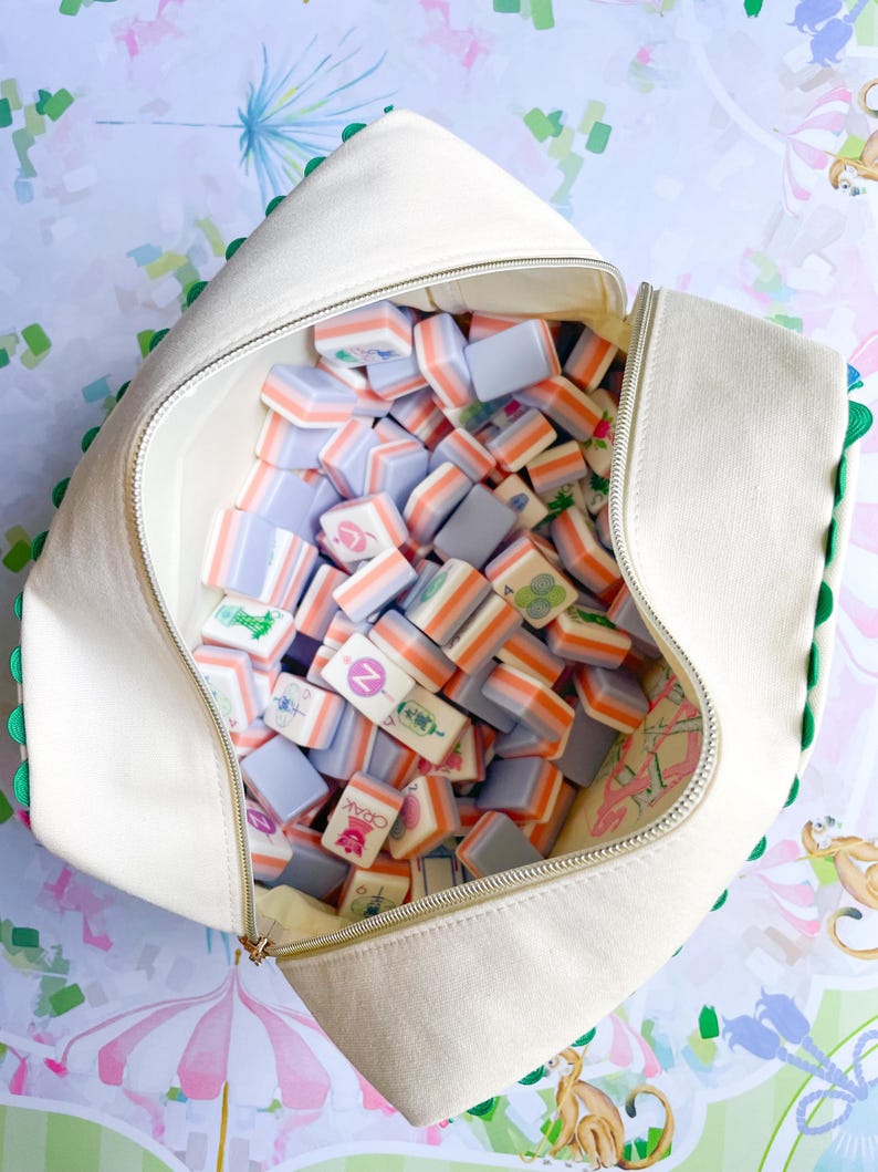 Puede incluir: Una bolsa de lona blanca, abierta y llena de coloridas fichas de Mahjong. Las fichas tienen una mezcla de colores, incluyendo naranja, lavanda y blanco. La bolsa tiene un ribete verde y una cremallera dorada. El fondo tiene un patr&oacute;n caprichoso.