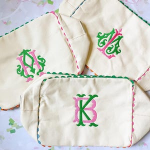 Puede incluir: Bolsas de lona blanquecinas con bordes festoneados en verde, rosa y azul. Cada bolsa presenta un monograma diferente en bordado verde y rosa. Las bolsas est&aacute;n dispuestas sobre una superficie estampada.