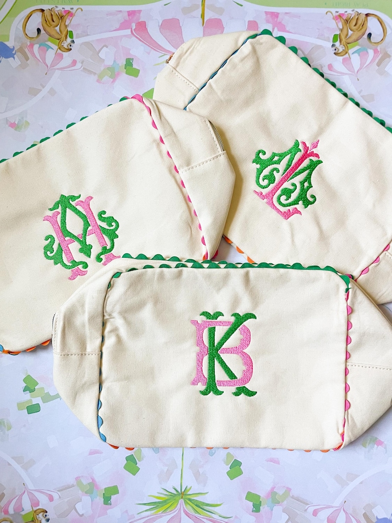 Puede incluir: Bolsas de lona blanquecinas con bordes festoneados en verde, rosa y azul. Cada bolsa presenta un monograma diferente en bordado verde y rosa. Las bolsas est&aacute;n dispuestas sobre una superficie estampada.