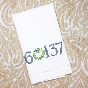 May include: White folded towel with embroidered blue numbers and a wreath design. The numbers "6" and "137" are embroidered in a dark blue thread, with a green wreath and blue bow detail.