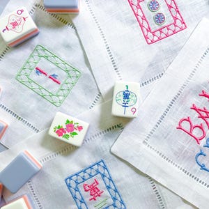 Puede incluir: Una colecci&oacute;n de fichas de Mahjong y servilletas de lino bordadas. Las fichas son blancas, azules y naranjas. Las servilletas presentan bordados coloridos en rosa, verde y azul, con costuras decorativas en los bordes. La escena es brillante y acogedora.
