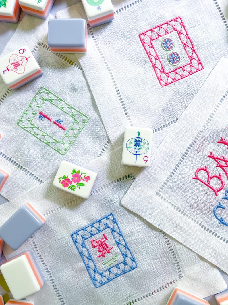 Puede incluir: Una colecci&oacute;n de fichas de Mahjong y servilletas de lino bordadas. Las fichas son blancas, azules y naranjas. Las servilletas presentan bordados coloridos en rosa, verde y azul, con costuras decorativas en los bordes. La escena es brillante y acogedora.