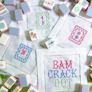 May include: A collection of Mahjong tiles with various symbols and colors, scattered around embroidered linen napkins. The napkins feature embroidered designs and the words "BAM CRACK DOT" in colorful thread. The tiles have a light blue top and orange and white sides.