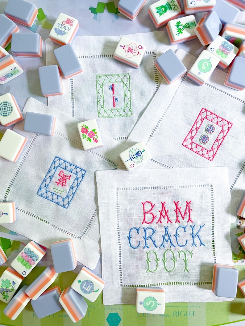 Puede incluir: Una colecci&oacute;n de fichas de Mahjong con varios s&iacute;mbolos y colores, dispersas alrededor de servilletas de lino bordadas. Las servilletas presentan dise&ntilde;os bordados y las palabras "BAM CRACK DOT" en hilo colorido. Las fichas tienen una parte superior azul claro y lados naranjas y blancos.