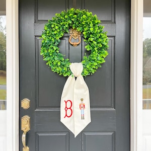May include: A green boxwood wreath hangs on a black door. A white banner with a red "B" and a nutcracker design is attached to the wreath. The door has a brass door knocker and handle.