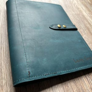 May include: Teal leather journal with a stitched spine and a flap closure secured by two brass-colored snaps. The journal has the embossed text "Lacey Gray" on the lower right corner. The leather has a distressed appearance.