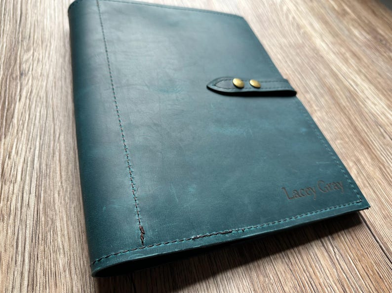 May include: Teal leather journal with a stitched spine and a flap closure secured by two brass-colored snaps. The journal has the embossed text "Lacey Gray" on the lower right corner. The leather has a distressed appearance.