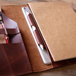 May include: A brown leather notebook cover with a built-in pocket for a tablet, pen, and other accessories. The cover is open to reveal a blank notebook and a tablet.
