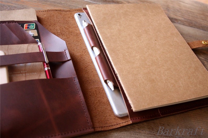 May include: A brown leather notebook cover with a built-in pocket for a tablet, pen, and other accessories. The cover is open to reveal a blank notebook and a tablet.