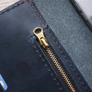 May include: A navy blue leather wallet with a gold zipper and brown stitching. The wallet is open, revealing a card slot and a pocket holding a phone and stylus. The leather has a distressed look.