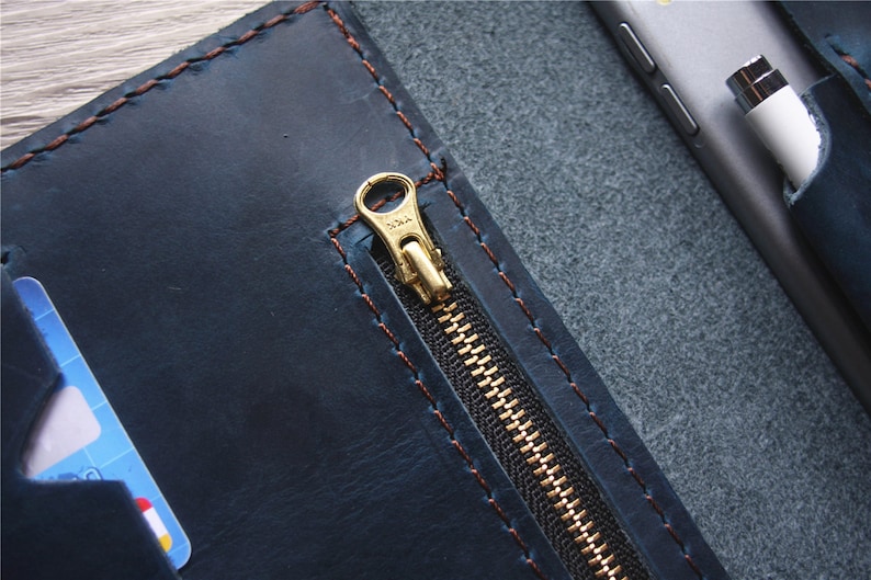 May include: A navy blue leather wallet with a gold zipper and brown stitching. The wallet is open, revealing a card slot and a pocket holding a phone and stylus. The leather has a distressed look.