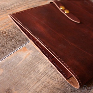 May include: A brown leather portfolio with a flap closure and brass accents. The portfolio is closed and sitting on a wooden surface.