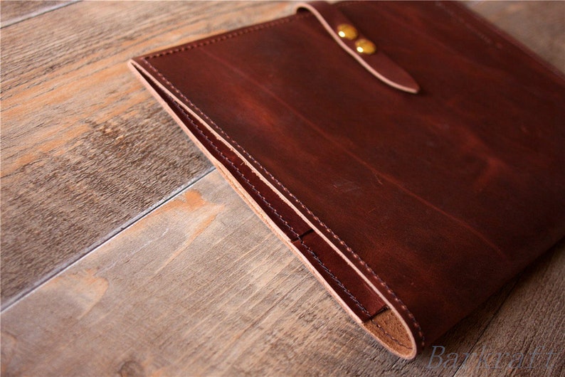 May include: A brown leather portfolio with a flap closure and brass accents. The portfolio is closed and sitting on a wooden surface.