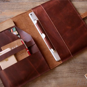 May include: A brown leather portfolio with multiple pockets and compartments. The portfolio is open and contains a tablet, a phone, a notebook, a pen, and a credit card.