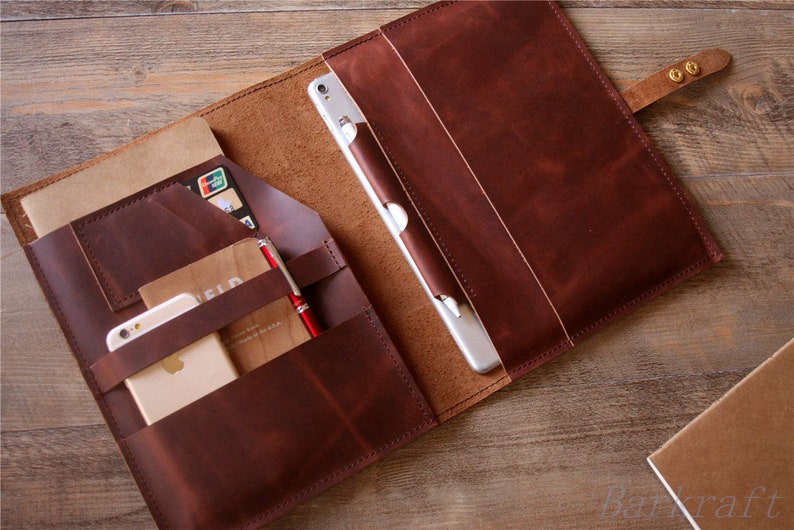 May include: A brown leather portfolio with multiple pockets and compartments. The portfolio is open and contains a tablet, a phone, a notebook, a pen, and a credit card.
