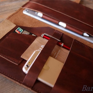 May include: A brown leather portfolio with multiple pockets and straps. The portfolio is open and contains a tablet, a phone, a pen, and credit cards.