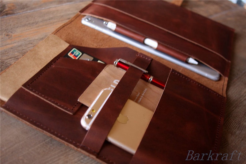 May include: A brown leather portfolio with multiple pockets and straps. The portfolio is open and contains a tablet, a phone, a pen, and credit cards.