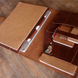 May include: A brown leather portfolio with a flap closure and multiple pockets. The portfolio is open and contains a tablet, a notebook, a pen, and a credit card. The portfolio is on a wooden surface.