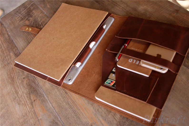 May include: A brown leather portfolio with a flap closure and multiple pockets. The portfolio is open and contains a tablet, a notebook, a pen, and a credit card. The portfolio is on a wooden surface.