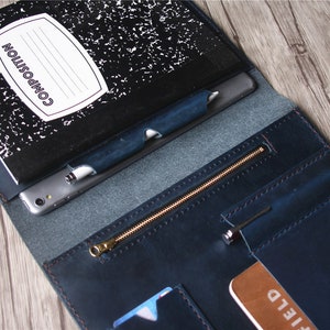 May include: Open leather folio in navy blue with a composition notebook, pen, and smartphone. The folio has a zippered pocket and card slots. The notebook is black and white with the word "COMPOSITION" on the cover.