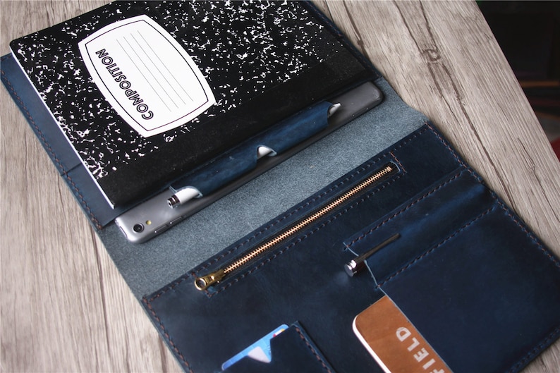 May include: Open leather folio in navy blue with a composition notebook, pen, and smartphone. The folio has a zippered pocket and card slots. The notebook is black and white with the word "COMPOSITION" on the cover.
