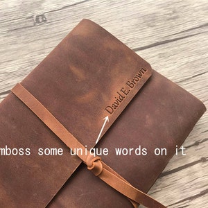 May include: A brown leather journal with a strap closure. The journal has the name "David E. Brown" embossed on the cover. The text "Emboss some unique words on it" is also visible.