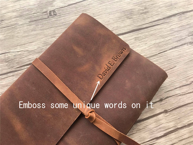 May include: A brown leather journal with a strap closure. The journal has the name "David E. Brown" embossed on the cover. The text "Emboss some unique words on it" is also visible.