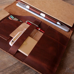 May include: A brown leather notebook cover with multiple pockets and slots for holding a phone, cards, and a pen. The cover is open to reveal a notebook with a brown paper cover.
