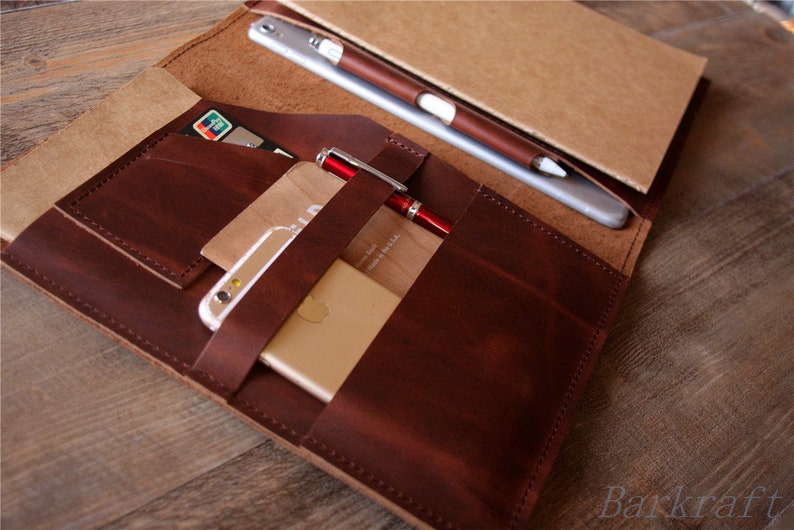 May include: A brown leather notebook cover with multiple pockets and slots for holding a phone, cards, and a pen. The cover is open to reveal a notebook with a brown paper cover.