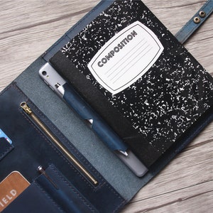 May include: Blue leather notebook cover with a composition notebook, smartphone, and pen. The cover has card slots, a zippered pocket, and a snap closure. The notebook has a white label that says "COMPOSITION".