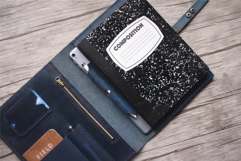 May include: Blue leather notebook cover with a composition notebook, smartphone, and pen. The cover has card slots, a zippered pocket, and a snap closure. The notebook has a white label that says "COMPOSITION".