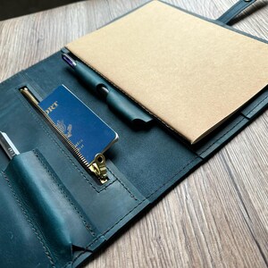 May include: A teal leather notebook cover with a passport, pen, and notepad. The cover has a zipper pocket and card slots. The notepad is tan, and the pen is black. The passport is blue with gold lettering.