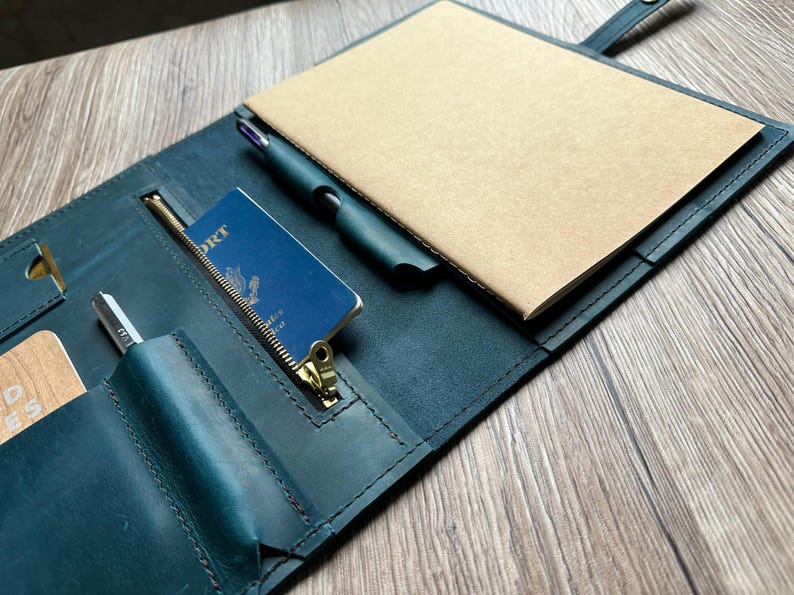 May include: A teal leather notebook cover with a passport, pen, and notepad. The cover has a zipper pocket and card slots. The notepad is tan, and the pen is black. The passport is blue with gold lettering.
