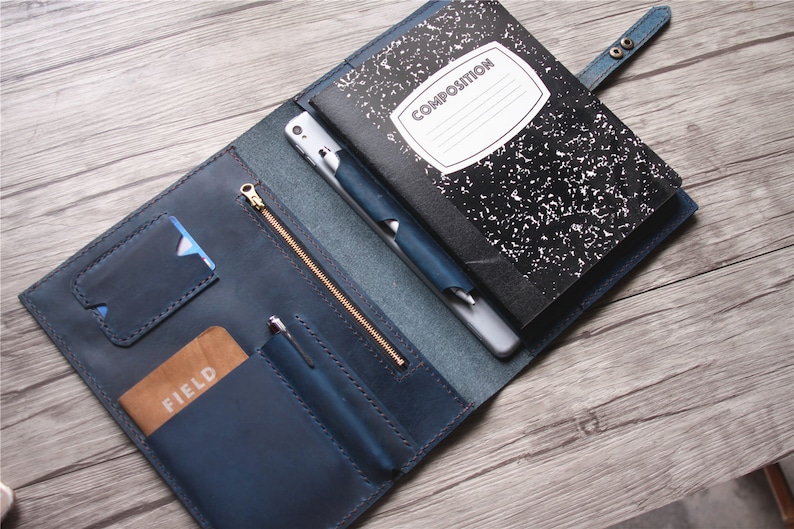 May include: Blue leather notebook cover with a gold zipper, card slots, and pen holder. A black and white composition notebook is inside, along with a pen and a phone. The cover is open, revealing the contents.