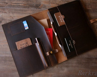 May include: Open brown leather portfolio with multiple pockets and slots. The portfolio contains a passport, a notebook, a pen, and a credit card. The text "FIELD NOTES" is visible on the notebook.