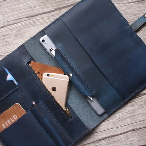 May include: A blue leather folio-style wallet, open to reveal compartments for a phone, cards, and a pen. The wallet has a snap closure and a zippered pocket. The interior includes a phone, a pen, and a card with the word "FIELD".