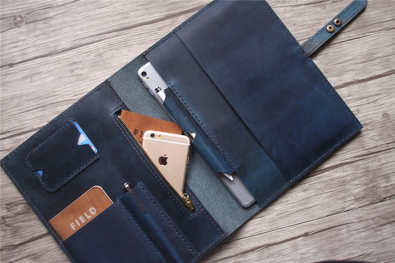 May include: A blue leather folio-style wallet, open to reveal compartments for a phone, cards, and a pen. The wallet has a snap closure and a zippered pocket. The interior includes a phone, a pen, and a card with the word "FIELD".
