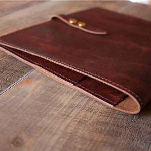 May include: A brown leather laptop sleeve with a flap closure and brass accents. The sleeve is folded in half, showing the interior lining.