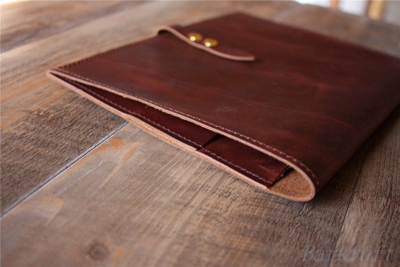 May include: A brown leather laptop sleeve with a flap closure and brass accents. The sleeve is folded in half, showing the interior lining.