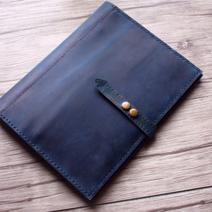 May include: A blue leather folio with a strap closure and two brass-colored button details. The edges are stitched with a contrasting red thread. The folio is on a wooden surface.