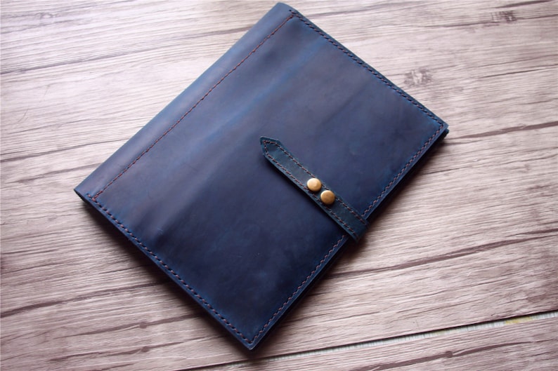 May include: A blue leather folio with a strap closure and two brass-colored button details. The edges are stitched with a contrasting red thread. The folio is on a wooden surface.