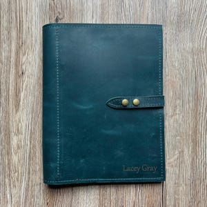 May include: A teal leather journal with a strap closure and brass-colored studs. The journal has stitched edges and the name "Lacey Gray" is embossed on the bottom right corner. The journal is on a wooden surface.